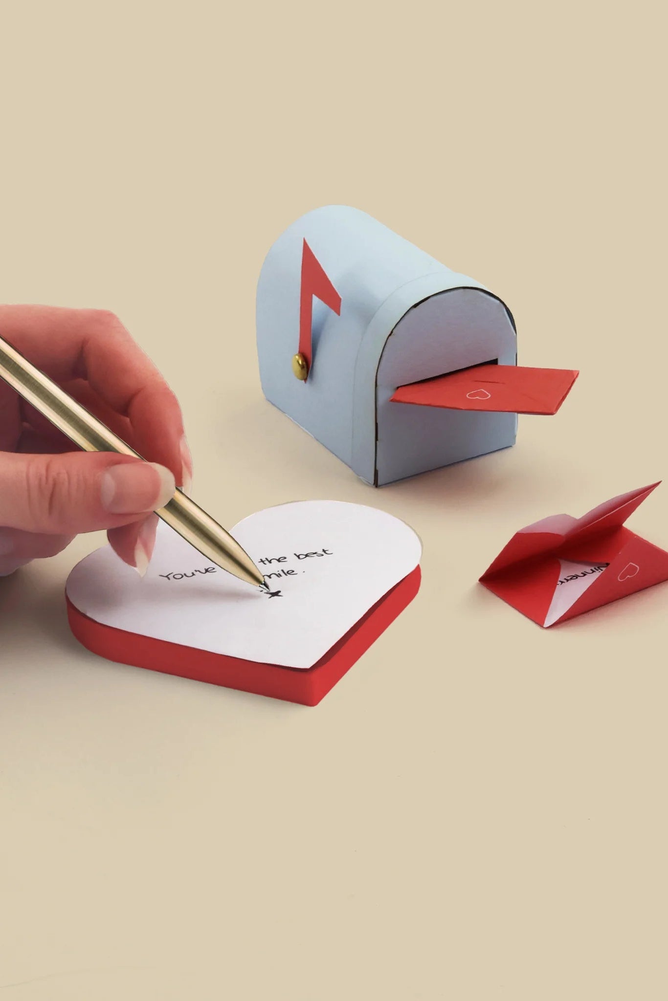 Hand writing on a heart-shaped notepad with a small mailbox and envelope in the background.