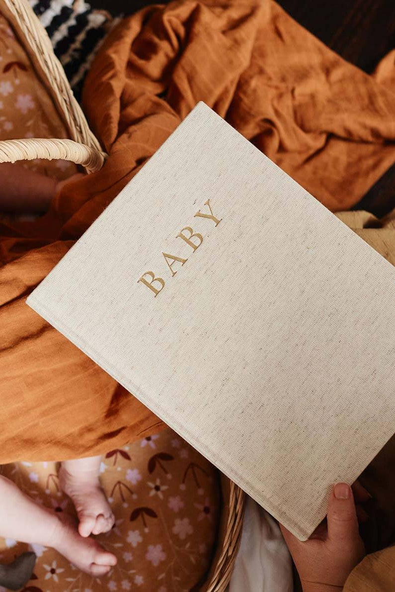 Beige book titled 'BABY' held by a hand on a textured surface with a basket and fabric.