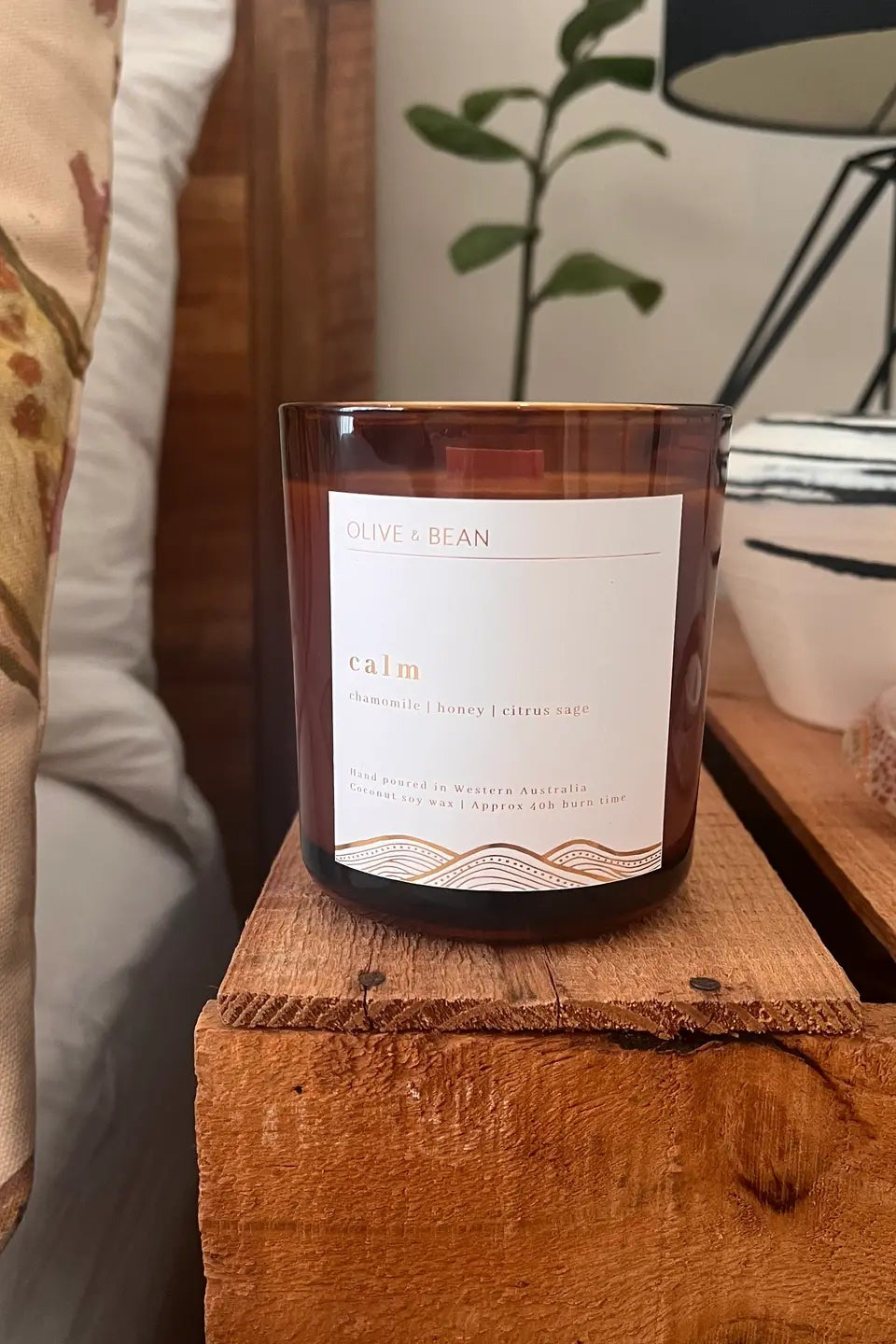Candle with a label on a wooden surface next to decorative pillows and a plant